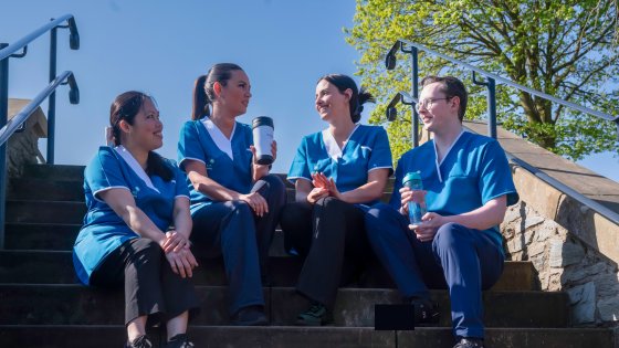 Four Leadcare Agency Nurses wearing blue scrubs sit together on outdoor steps, smiling and chatting while holding reusable coffee cups. The scene is bright and sunny with a clear blue sky and green leafy trees in the background in Northern Ireland.