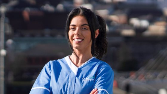 A smiling woman wearing a blue Leadcare Agency Nurse uniform stands outdoors with arms crossed. She has dark hair pulled back in a ponytail, and the background shows a blurred cityscape in Northern Ireland.